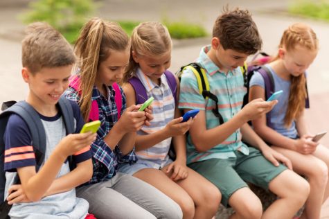 65551235 - primary education, friendship, childhood, technology and people concept - group of happy elementary school students with smartphones and backpacks sitting on bench outdoors
